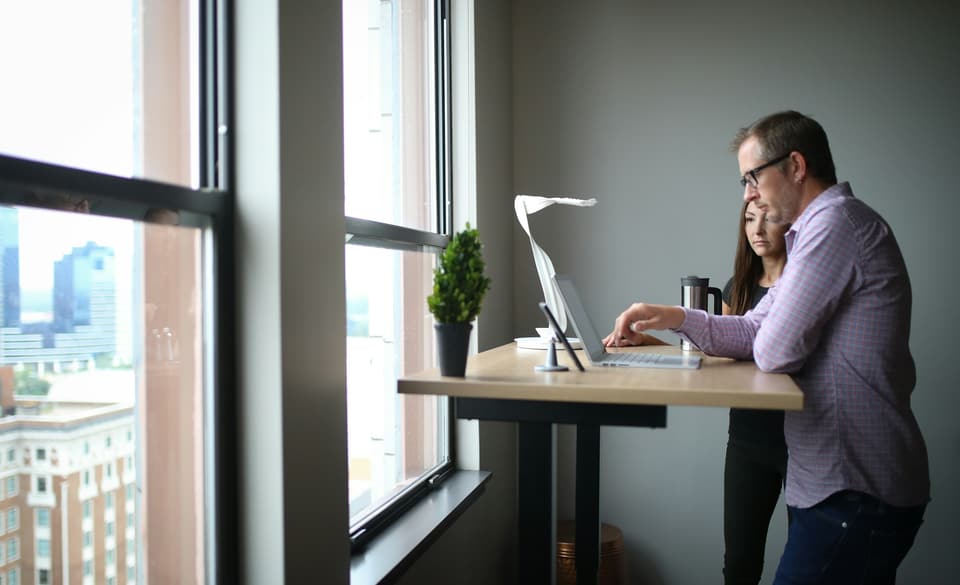 Two people at a standing desk, looking at a laptop by a window